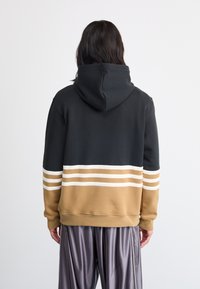 Hooded sweatshirt ima črno zgornjo in peščeno spodnjo del ter bele vodoravne črte. Material se zdi mehak in ima sproščen kroj.