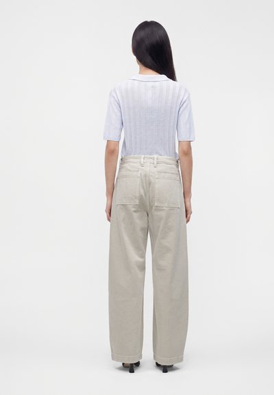 Femme aux longs cheveux foncés portant un haut bleu clair à manches courtes et un pantalon beige à larges jambes, debout de dos devant un fond uni.