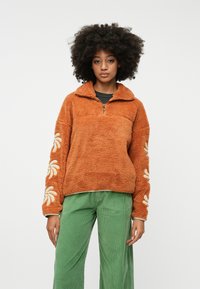 Fleeceový pulóver v spálenej oranžovej farbe s polovičným zipsom, zdobený smotanovým kvetinovým vyšívaním na rukávoch. Spárovaný so zelenými kordovými nohavicami.