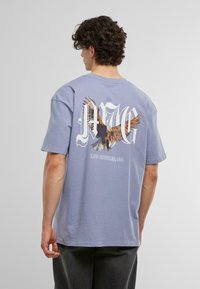 Tricou din bumbac de culoare albastru deschis, cu o croială lejeră, având un grafic cu un vultur și textul "LOS ANGELES, CA" scris cu alb pe spate. Mâneci scurte.