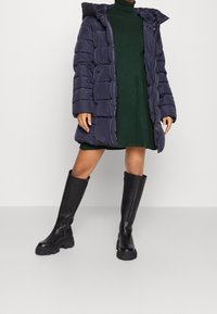Manteau matelassé bleu marine avec capuche, présentant des coutures horizontales et une coupe mi-longue, porté par-dessus une robe verte et assorti à des bottes noires montantes jusqu'au genou.