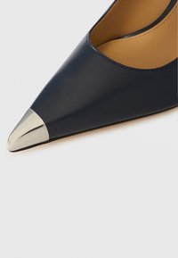 Mørkeblå lærhøyhæl med spiss tå og metallisk sølvhette. Glatt tekstur, elegant design og minimale sømmer.