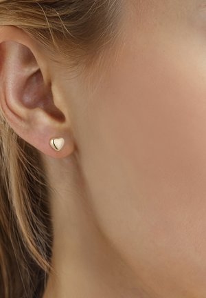 Primer plano de una oreja izquierda que lleva un pequeño pendiente de botón en forma de corazón dorado y brillante, sobre piel clara y cabello castaño claro.