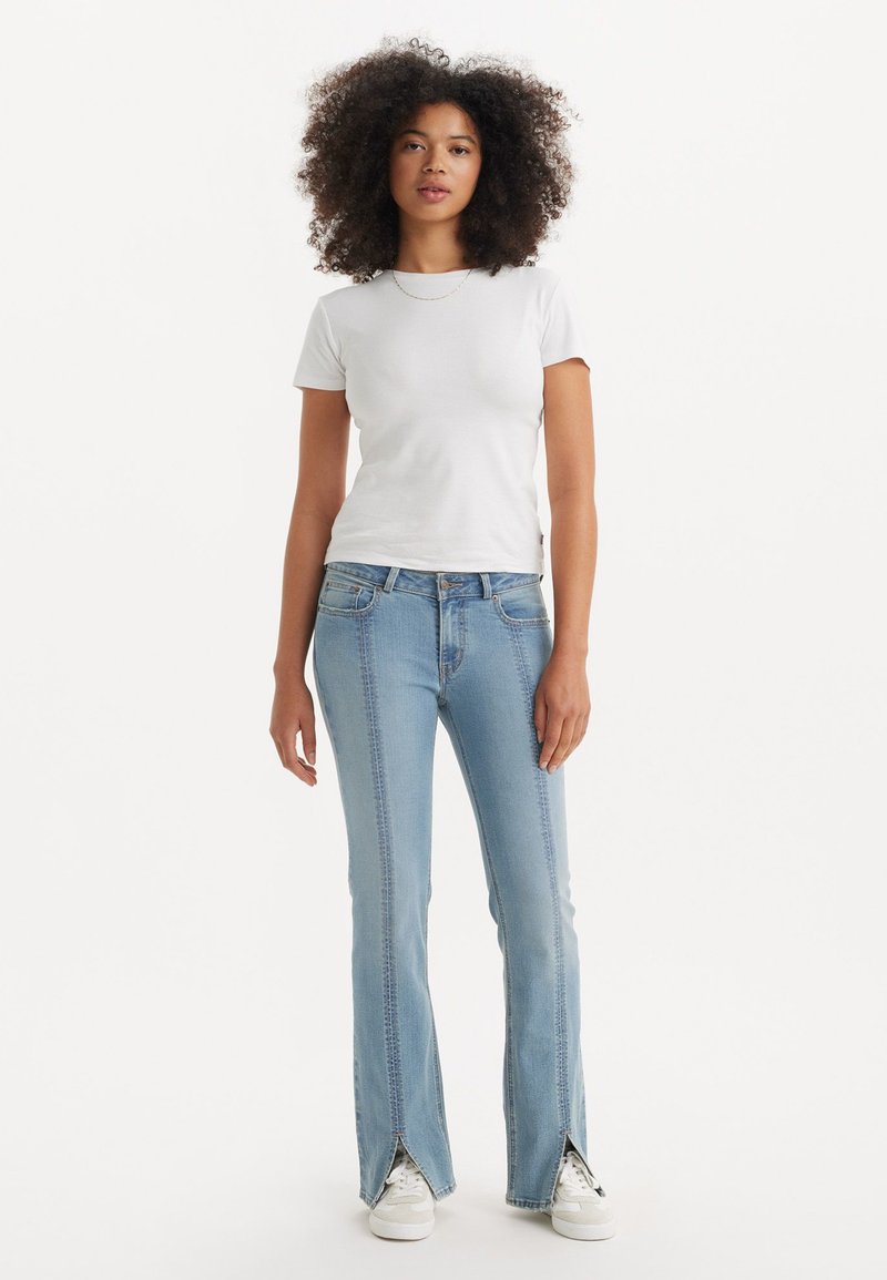 Jean évasé bleu clair avec un design à fente à l'avant. Associé à un t-shirt blanc à manches courtes et des baskets blanches. Tenue simple et décontractée.