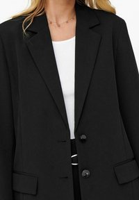 Nainen, jolla on päällä musta muotoiltu bleiseri valkoisen ribattun topin päällä, ja asusteena herkät korut, joissa on pieniä helmiä.