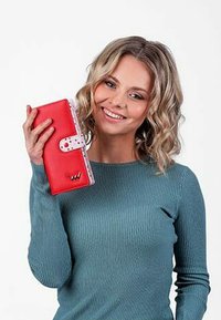 Červená peněženka vyrobená z hladkého materiálu s puntíkovanými akcenty, s uzavřením na patentku a raženým logem, kterou drží osoba v modrém svetru.