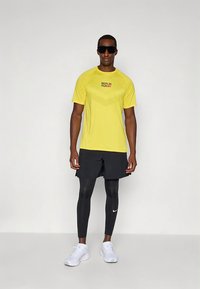 Gul kortermet løpetrøye med teksten "BERLIN RUN", kombinert med svarte shorts og svarte leggings. Hvite joggesko fullfører antrekket.