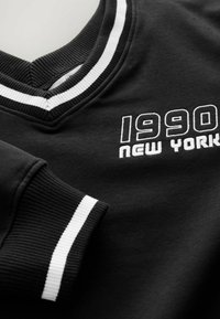 Černá mikina s výstřihem do V a bílým pruhovaným ribkovaným límcem. Na přední straně je bílé vyšívané označení "1990 NEW YORK".
