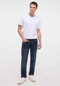 Conjunto masculino que presenta una camiseta de algodón blanca y jeans de mezclilla azul oscuro, complementado con zapatillas blancas con detalles en marrón. Diseño sencillo y ajuste casual.