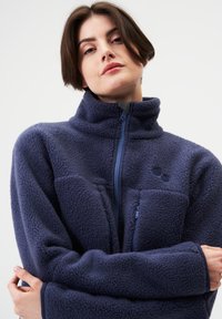 Marineblauer Fleecepullover mit hohem Kragen, Frontreißverschluss und Brusttasche; zeichnet sich durch ein strukturiertes, weiches Gewebe und dezente Logodetails aus.