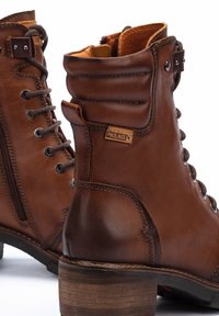 Boots en cuir marron à la cheville avec des lacets et une fermeture éclair sur le côté, présentant des textures contrastées, un talon empilé et une petite étiquette logo sur le côté.