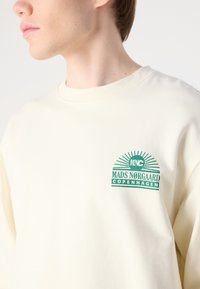 Felpa color crema con collo rotondo. Presenta un logo grafico verde con la scritta "MADS NØRGAARD COPENHAGEN" sul lato sinistro del petto. Texture morbida.