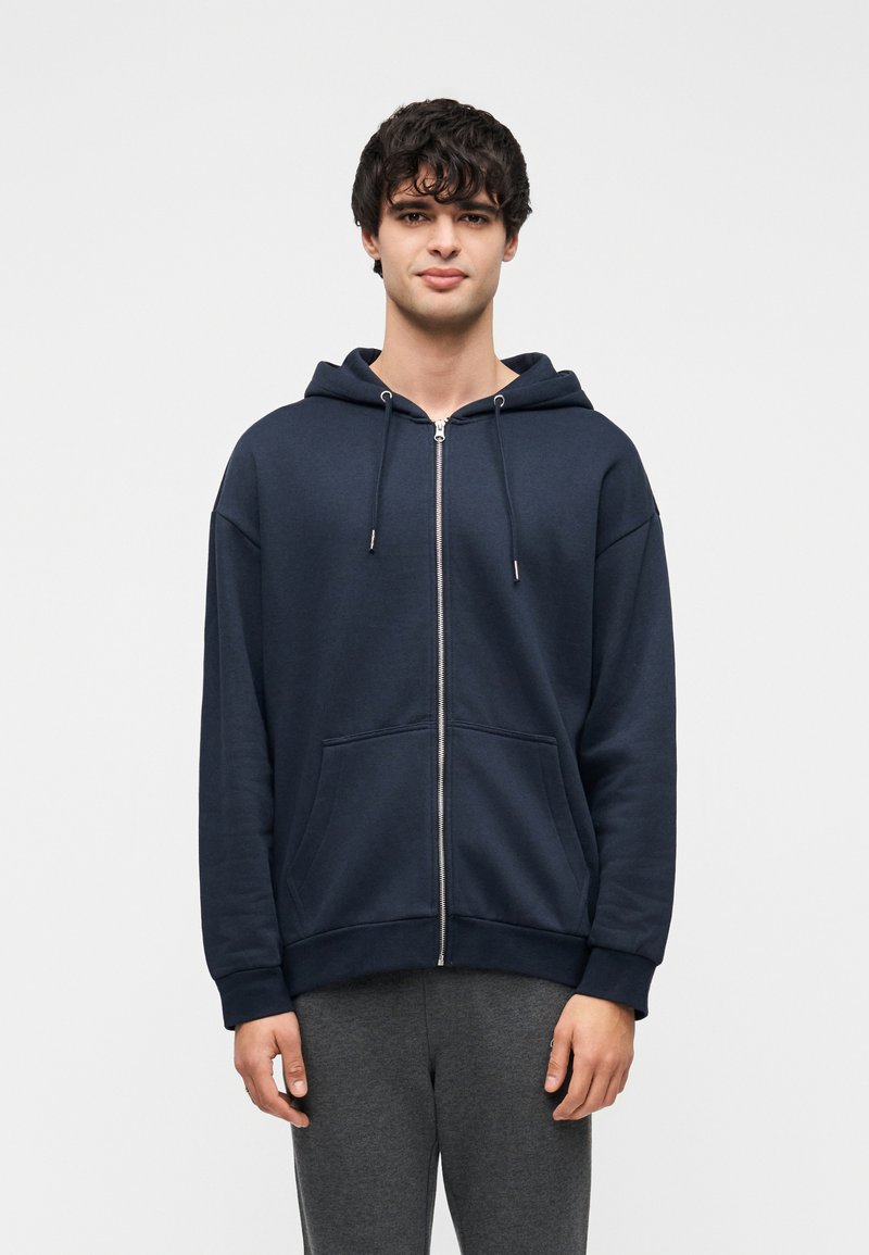 Σκούρο μπλε φούτερ με φερμουάρ, μπροστινή τσέπη τύπου καγκουρό, κουκο ύλα με κορδόνι και ριμπ μανσέτες. Μαλακή υφή, άνετη εφαρμογή και μίνιμαλ σχεδιασμός.