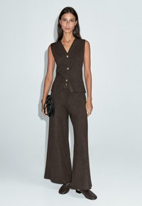 Femme portant un gilet marron sans manches à boutons, un pantalon large assorti, des chaussures marron, tenant un sac à main noir sur un fond uni.