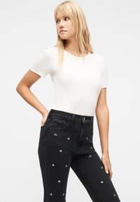 Γυναίκα με μακριά ξανθά μαλλιά που φοράει λευκό εφαρμοστό μπλουζάκι με κοντά μανίκια και μαύρο τζιν ψηλόμεσο με μικρές λευκές κεντημένες μέλισσες.