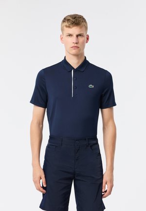 Marinblå pikétröja i mjukt tyg med en treknapplösning och kontrasterande vit kant. Har en grön broderad logotyp på bröstet.