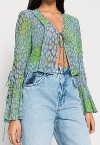 Cropped, sheer blouse in green and blue with a playful animal print. Features a V-neck, tie detail, and flared sleeves. Paired with light denim.