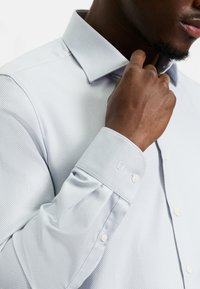 Hellblaues Hemd mit einer strukturierten Musterung, einem strukturierten Kragen und Knopfmanschetten. Verfügt über einen einzelnen Knopf an der Manschette und eine taillierte Passform.