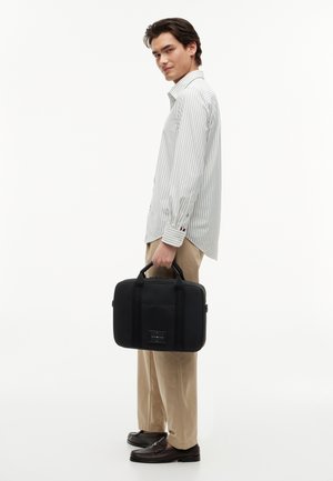 Joven con camisa de rayas y pantalones beige sosteniendo un maletín negro, de pie de lado contra un fondo blanco.