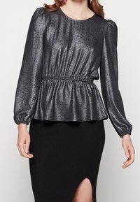 Top peplum noir brillant avec de longues manches bouffantes et une taille élastique ; présente une texture lisse et un léger éclat. Associé à une jupe noire côtelée.