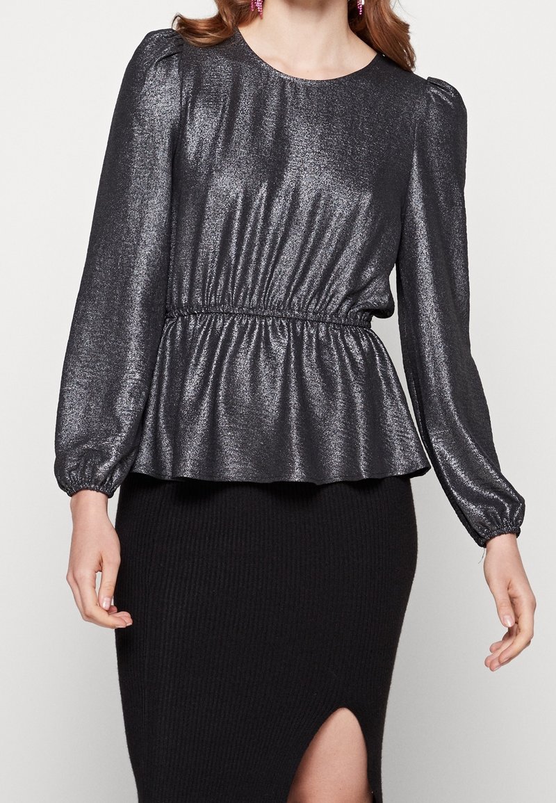 Top peplum noir brillant avec de longues manches bouffantes et une taille élastique ; présente une texture lisse et un léger éclat. Associé à une jupe noire côtelée.