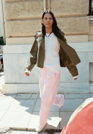Jeune femme marchant à l'extérieur portant une veste verte par-dessus une chemise blanche, un pantalon rose clair et des chaussures blanches, devant un bâtiment en pierre.