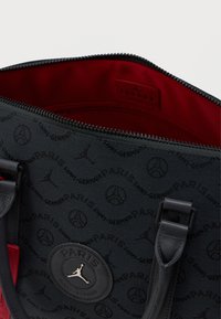 Fekete szövet táska a Paris Saint-Germain és Jumpman logókkal, fekete pántokkal és piros belső béléssel, Jordan címkével a belsejében.