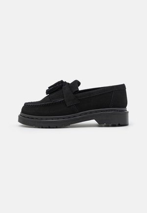 Mocassin en daim noir avec détail pompon, semelle épaisse et talon bas, présenté en profil latéral sur fond blanc.