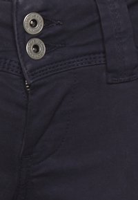 Emбaxas de ganga azul-marinho escuras exibem uma textura suave, com dois fechos de botão de metal e costura visível ao longo da cintura e dos bolsos.