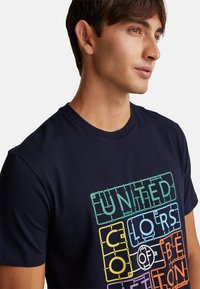 Námořnická modrá tričko vyrobené z bavlny s barevným grafickým potiskem a textem "UNITED COLORS OF BENETTON" v různých výrazných barvách.