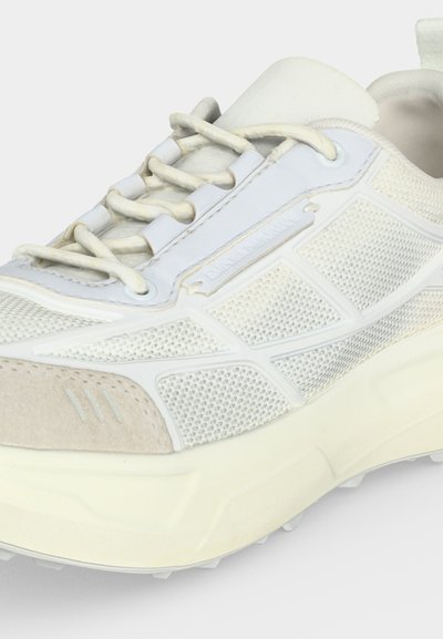 Baskets blanches de sport avec tissu en mesh, bout en daim, semelle épaisse et lacets blancs passés dans des œillets renforcés.