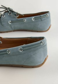 Chaussures bateau en daim bleu clair avec des lacets en cuir passés à travers des œillets métalliques et des semelles en caoutchouc marron clair, montrées sous des angles latéraux et arrière.