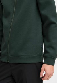 Dark green zip-up sweatshirt made of smooth, soft fabric; features a silver zipper, ribbed cuffs, and a small brand label on the side.