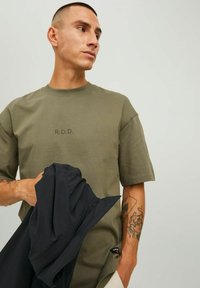 T-shirt verde oliva a maniche corte con stampa 'R.D.D.' sul petto. Presenta un collo rotondo, vestibilità comoda e un'etichetta sull'orlo. Tiene un indumento nero.