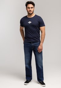 Młody mężczyzna stoi, ubrany w granatowy t-shirt z logo, niebieskie dżinsy i czarne trampki na jednolitym jasnoszarym tle.