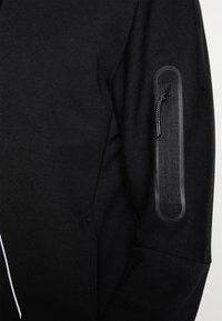 Sudadera negra con un tejido texturizado. Presenta un bolsillo con cremallera en la manga y acentos en negro contrastante a lo largo de la cremallera.