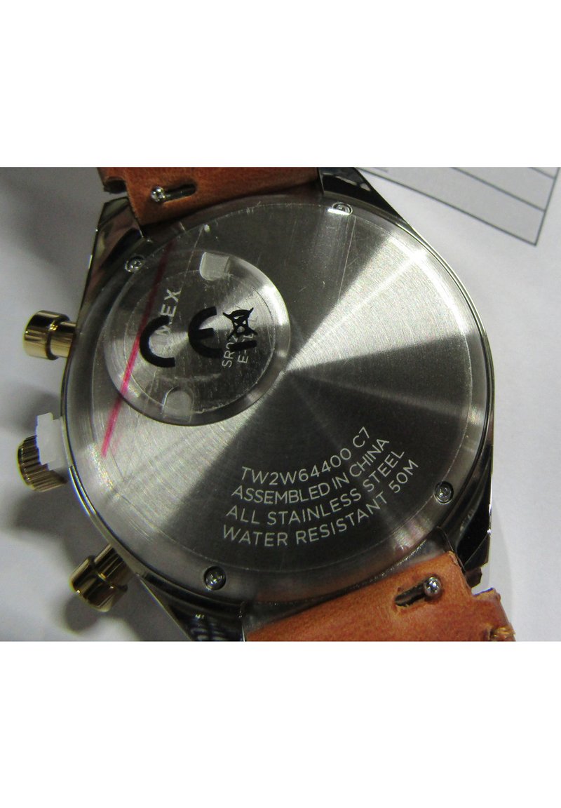 Boîtier de montre en acier inoxydable avec un bracelet en cuir marron. L'arrière présente un texte gravé sur l'assemblage, les matériaux et la résistance à l'eau.