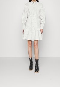 Robe en coton blanc avec une taille cintrée et une jupe évasée, dotée de fermetures à boutons et de détails perforés. Portée avec des bottines noires.