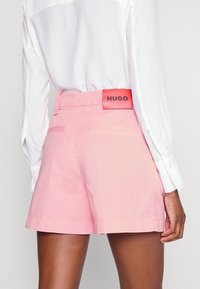 Lyserøde shorts lavet af letvægtsstof har en glat tekstur, for- og baglommer samt en mærket rød detalje på kanten.