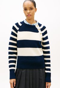 Μπλε ναυτικό και κρεμ ριγέ πλεκτό πουλόβερ με στρογγυλή λαιμόκοψη, ριμπ τελείωμα και κουμπιά στον ώμο. Το ύφασμα φαίνεται υφασμένο και με υφή.