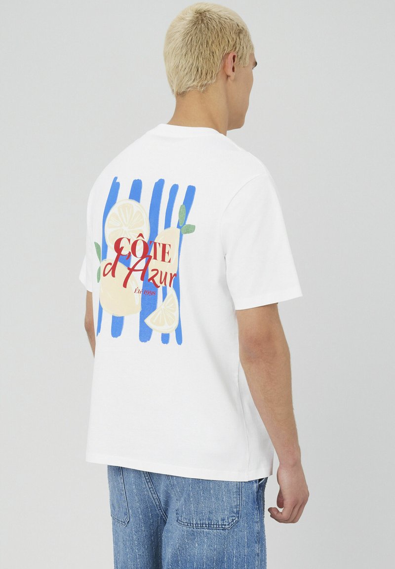 T-shirt en coton blanc avec un design graphique coloré, des rayures bleues, des illustrations de citrons et le texte "CÔTE d'Azur" en rouge.