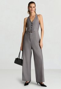 Donna che indossa un gilet grigio senza maniche con bottoni, pantaloni a gamba larga abbinati, tacchi neri, orecchini a cerchio argento e tiene una borsa nera.