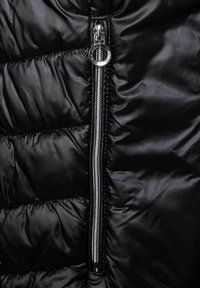 Veste matelassée noire avec une fermeture éclair argentée bien visible sur le devant, texture lisse et détails de couture matelassée.