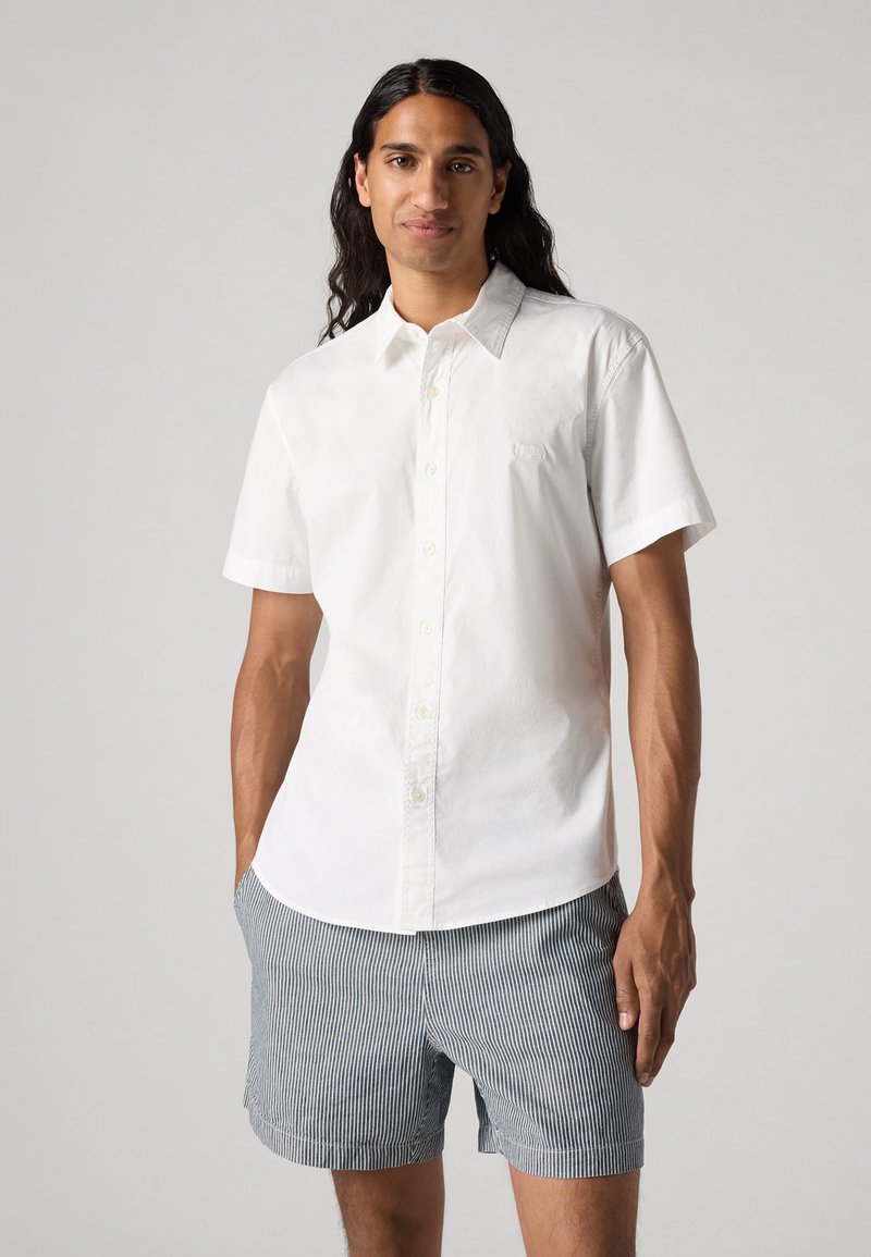 Chemise blanche à manches courtes avec un col boutonné, arborant un logo discret. Associée à un short rayé bleu et blanc, elle présente un look décontracté.