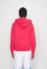 Felpa rossa con cappuccio e coulisse, maniche a costine. Presenta una texture liscia e una tasca a marsupio. Indossata con pantaloni bianchi con dettagli in tie-dye blu.