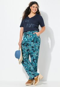 T-shirt V en marine avec "RIDE THE WAVE" en gris, associé à un pantalon à motif feuillage teal et des sandales compensées marron. Tenant un chapeau en paille bleu.