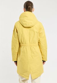 Veste jaune jusqu'aux genoux avec capuche, taille élastique et poches latérales. Fabriquée en matériau lisse et résistant à l'eau. Le dos présente un détail d'ourlet fendu.