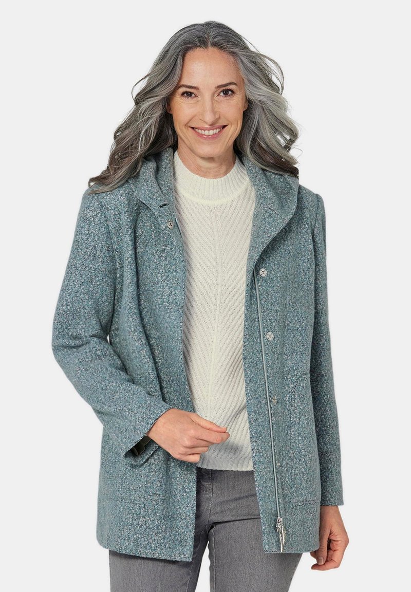 Blau strukturierter Mantel mit umgeschlagenem Kragen, Druckknöpfen und Seitentaschen, über einem cremefarbenen Strickpullover und grauen Jeans getragen.