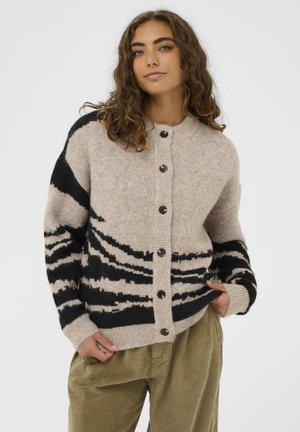 Young woman with curly hair wearing a beige buttoned cardigan with black abstract stripes and olive green corduroy pants.