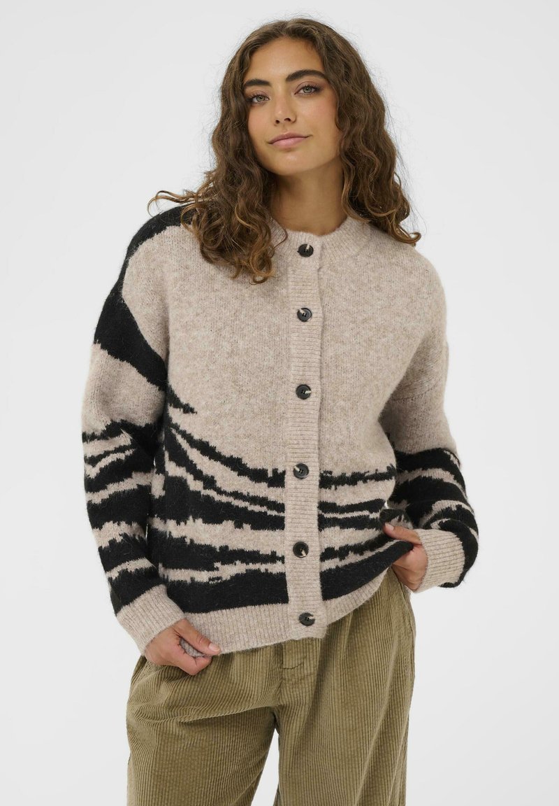 Mlada ženska z kodrastimi lasmi, oblečena v bež pulover s gumbi in črnimi abstraktnimi črtami ter olivno zelenimi kordna hlačami.